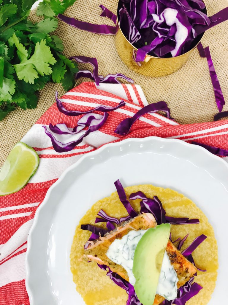 Easy, spice-rubbed salmon tacos with coconut-lime crema and avocado on a white plate, with cilantro, lime, and red cabbage in the background. 