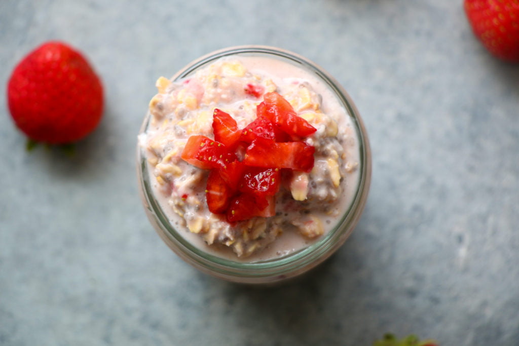 Strawberries and cream overnight oats in a glass cup, topped with fresh strawberries, on a gray background.