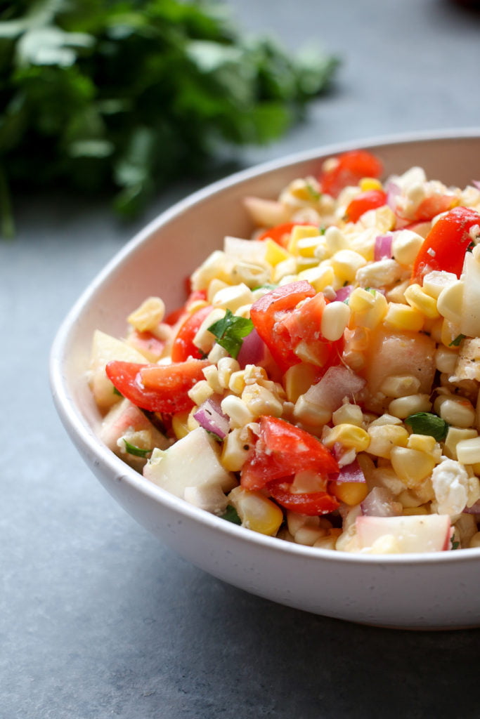Summer corn salad with stone fruit and feta, in an off-white bowl, on a gray background, up-close.