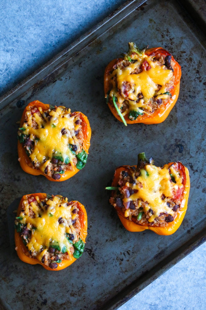4 vegetarian quinoa-stuffed peppers, topped with cheese, on a black baking sheet.