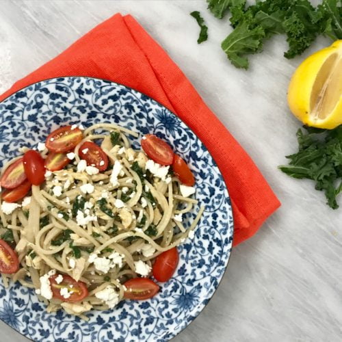 Kale, chicken and hummus pasta in a blue floral bowl, on a red towel, on a marble background.