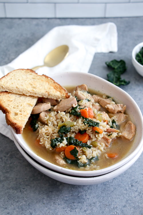 Chicken, kale and rice soup in a white bowl with toast on the side, on a gray background. 