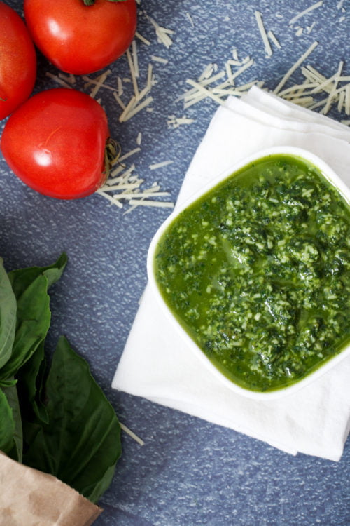 Basil pesto in a small white bowl, on a white towel and a blue background.
