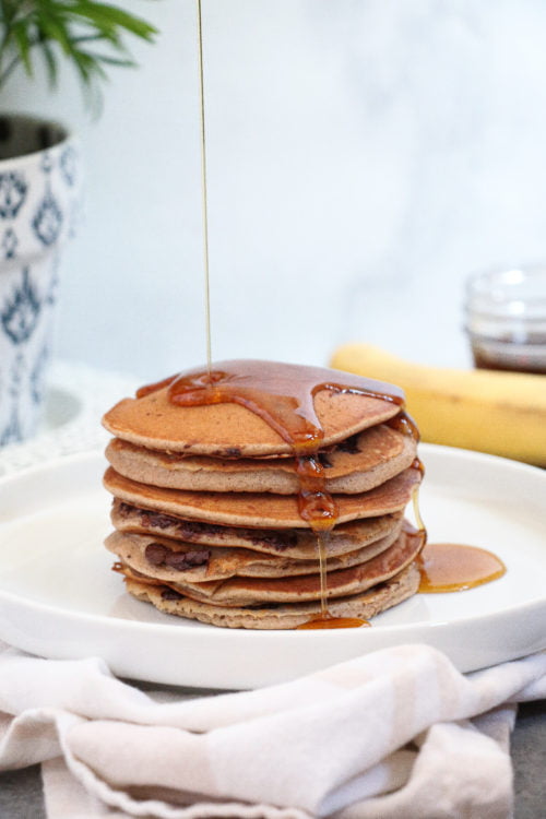 Stack of chocolate chip-banana pancakes drizzled with maple syrup on a white plate