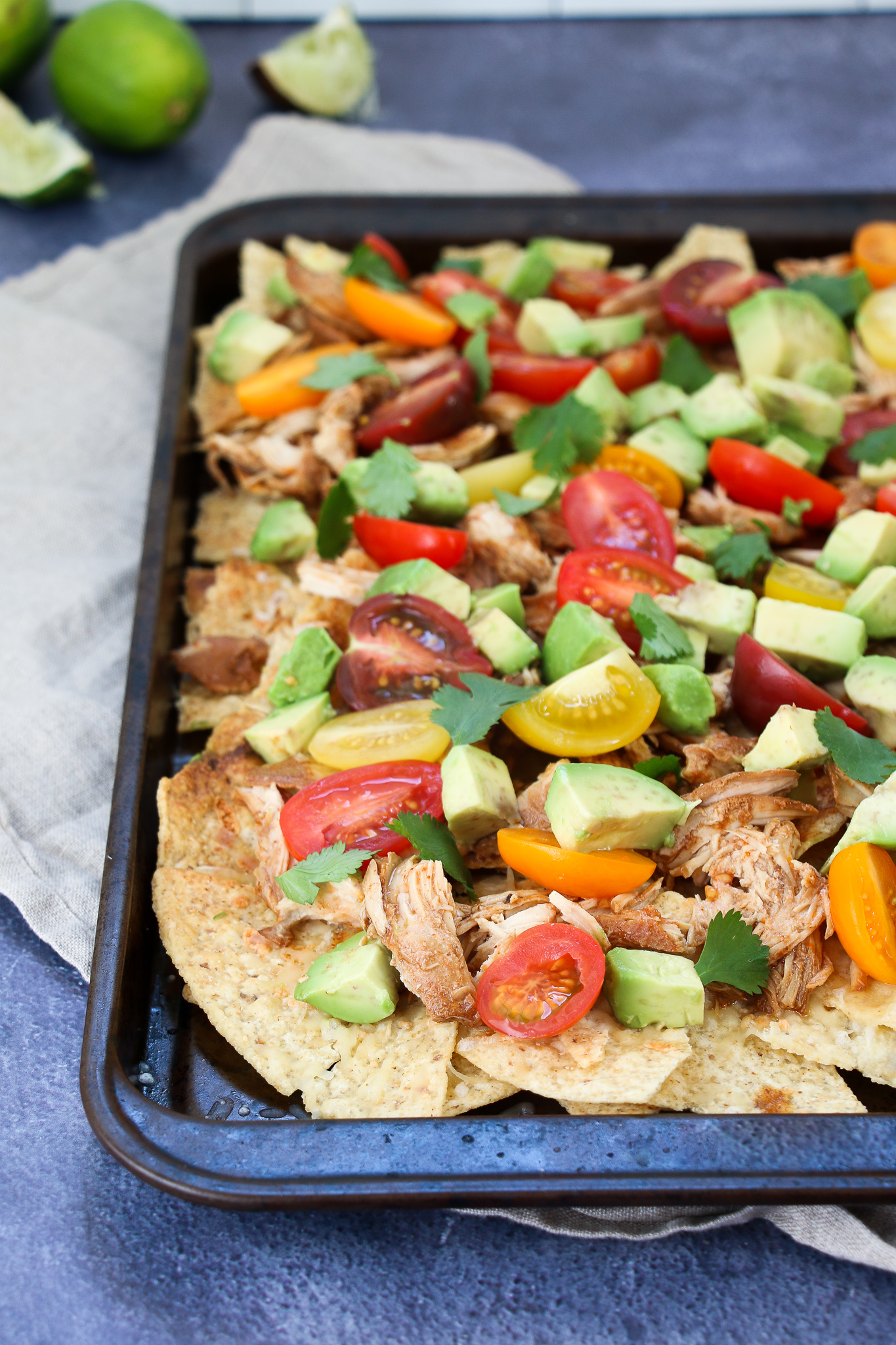 Healthy chicken nachos chopped with cherry tomatoes, and avocado on a black sheet pan, on a dark blue background.
