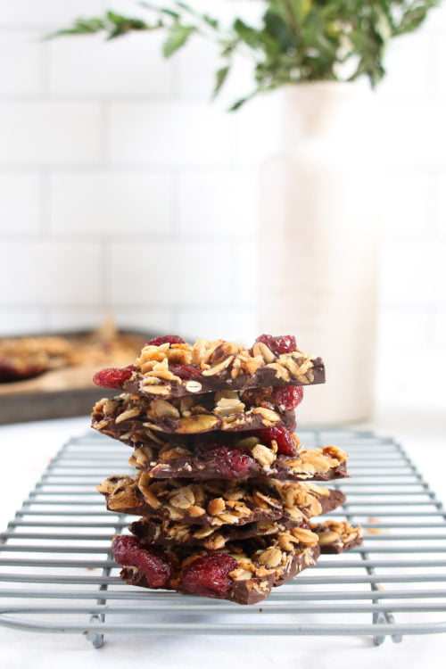 Dark chocolate granola bark pieces stack on top of each other, on a cooling rack on a white background