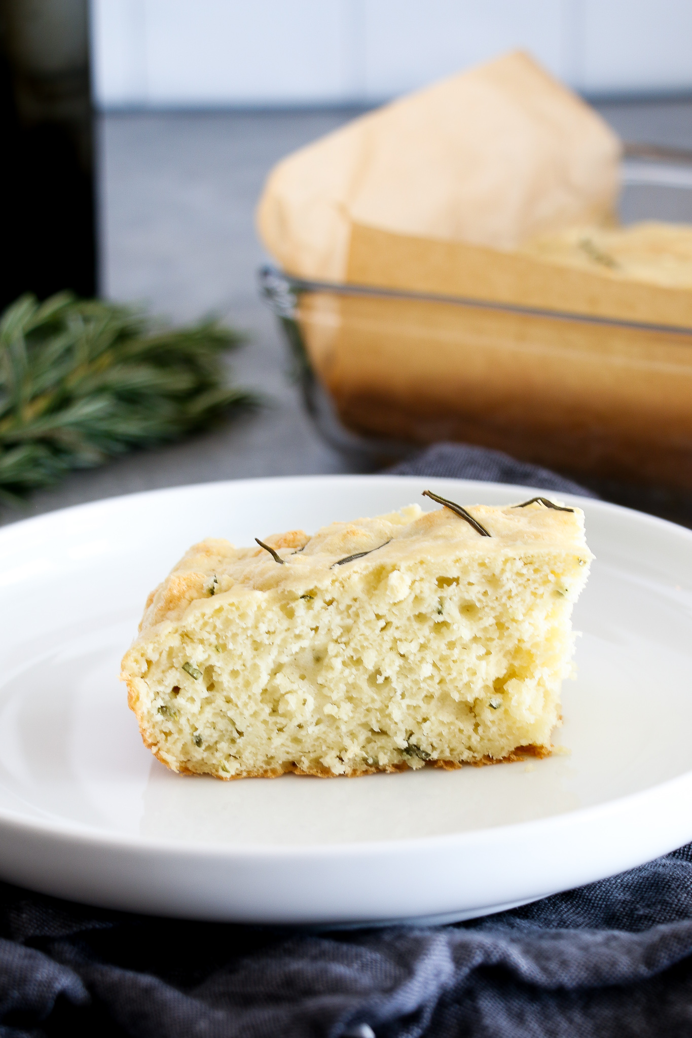 A slice of gluten-free rosemary and olive oil focaccia on a white plate on a blue background.