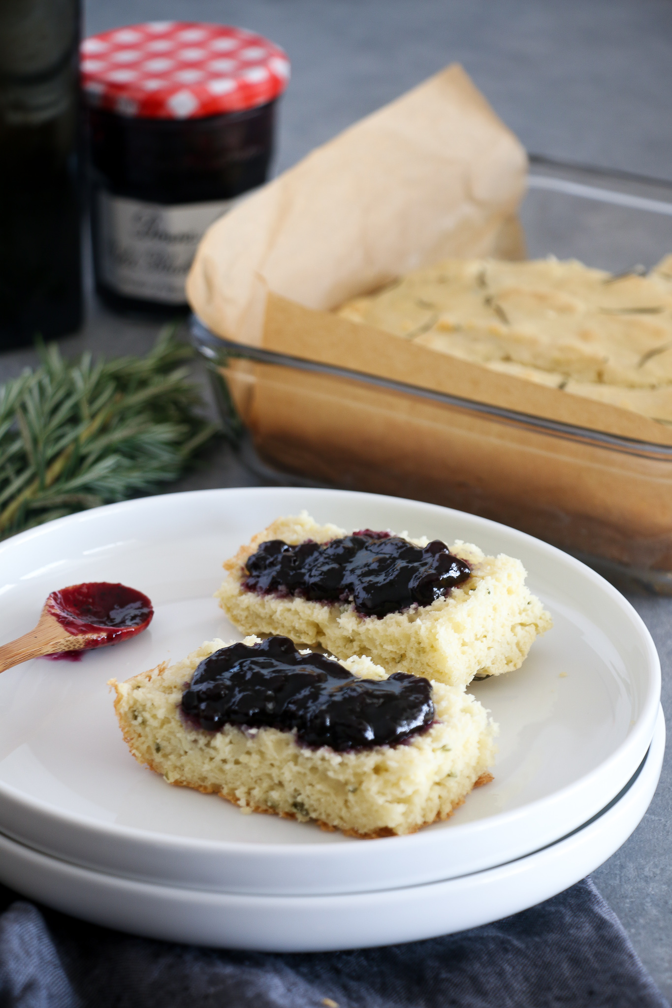Gluten-free rosemary and olive oil focaccia, topped with blueberry jam, on a white plate, on a gray background.