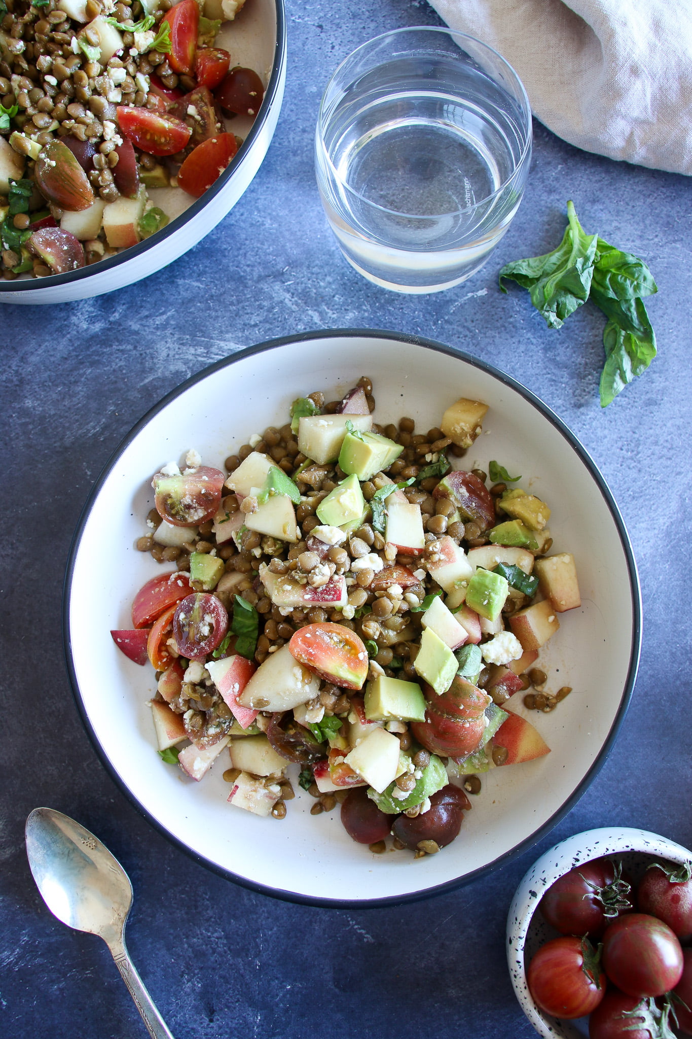 Cold lentil salad with peaches and feta in a white bowl on a blue background with a glass of water, and cherry tomatoes